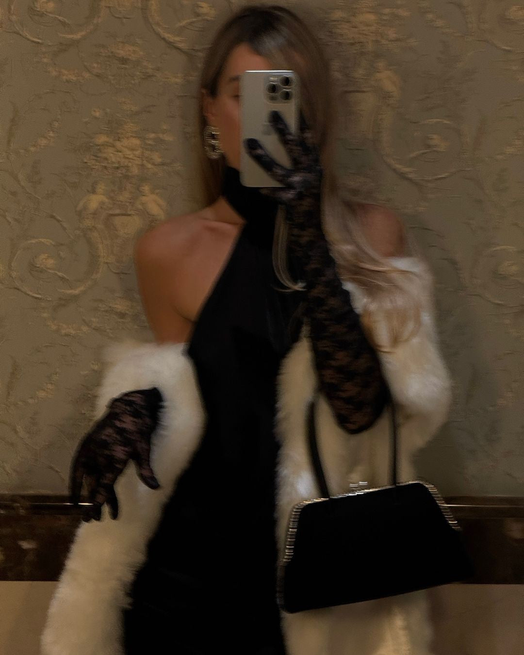 A person in a black halter-neck dress, long black lace gloves, and a white fur coat takes a mirror selfie with a phone, holding a black clutch bag in the other hand.