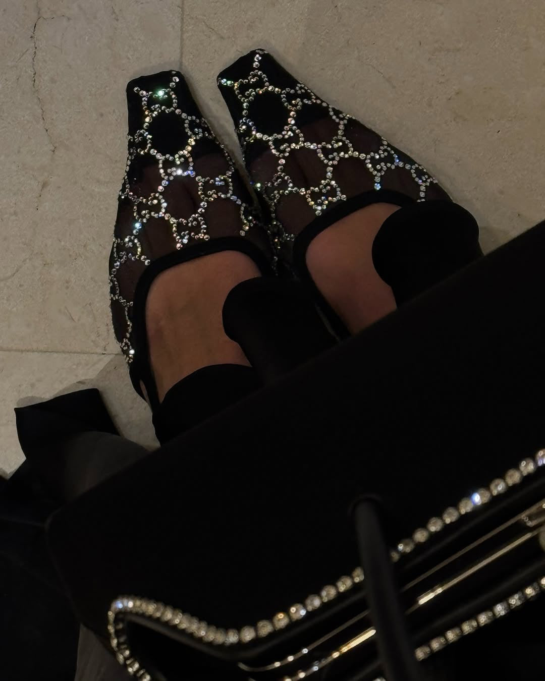 A close-up, high-angle shot of a person's feet wearing black mesh slingback pumps with crystal embellishments, likely Gucci, with a dark, possibly black, bag or garment partially visible in the foreground.