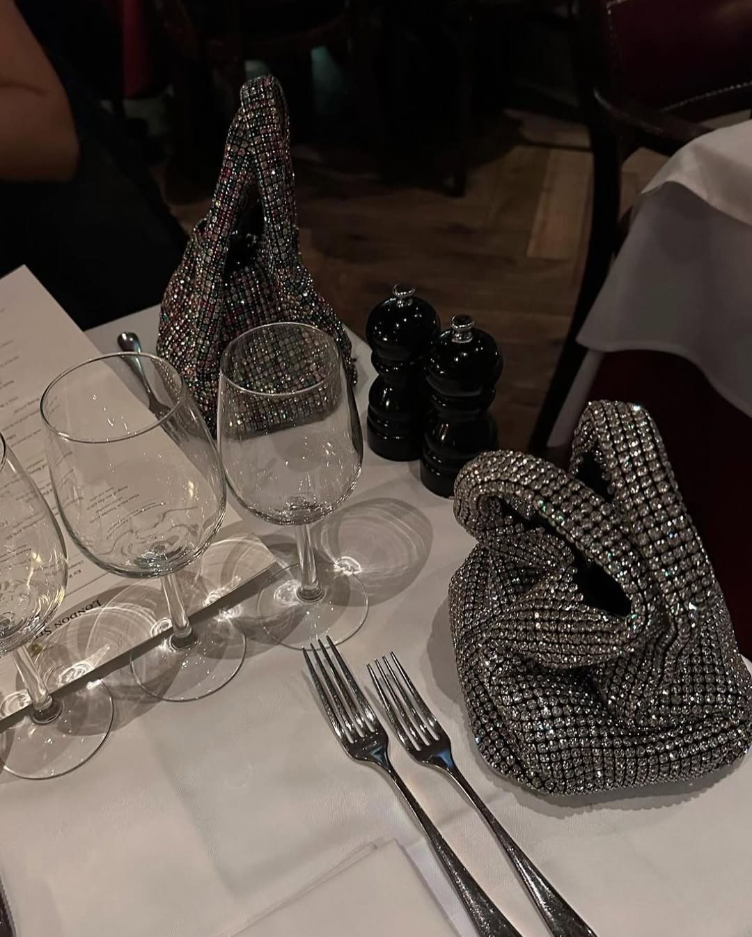 A close-up shot of a dining table set with two wine glasses, two forks, and two sparkling, rhinestone-covered handbags. A black salt and pepper shaker set is also visible on the table.