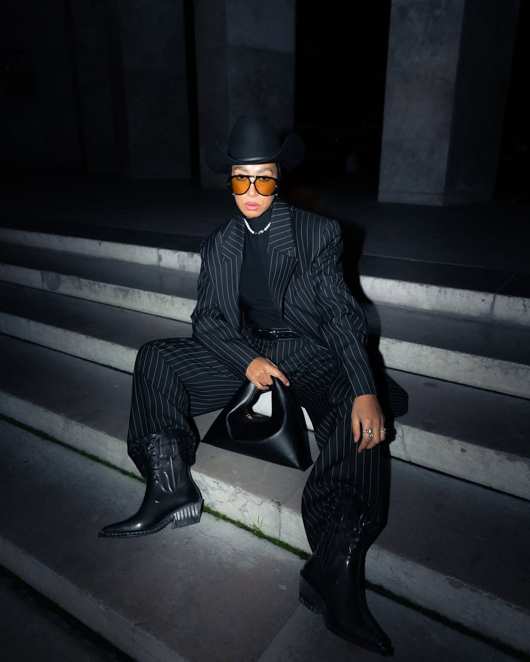 A person wearing a black pinstripe suit, a black turtleneck, a black cowboy hat, and black cowboy boots sits on concrete steps at night. They are holding a black structured handbag and wearing orange-tinted sunglasses and a pearl necklace. 