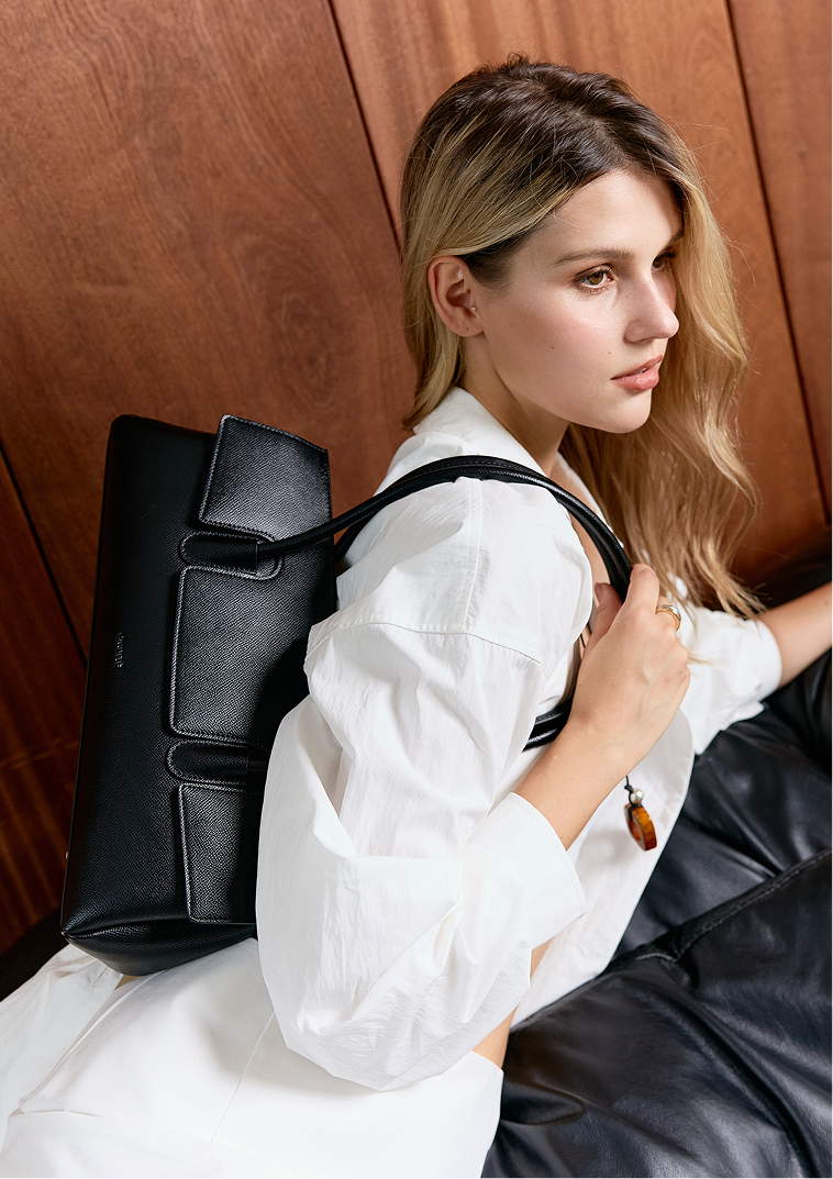 A woman with blonde hair wearing a white shirt and holding a black handbag over her shoulder, looking to the right. 