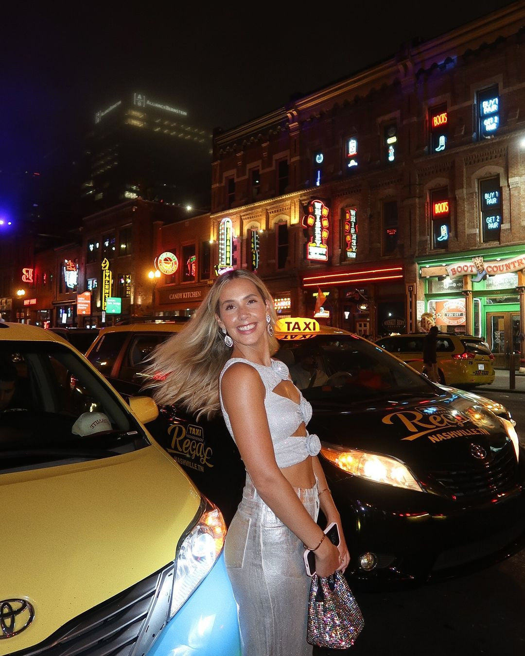 A smiling woman with blonde hair, wearing a silver crop top and matching skirt, stands on a city street at night. Taxis are parked nearby, and the background features buildings illuminated by numerous colorful neon signs, including one that says TAXI and another BOOTS COUNTR. The scene suggests a lively urban environment.