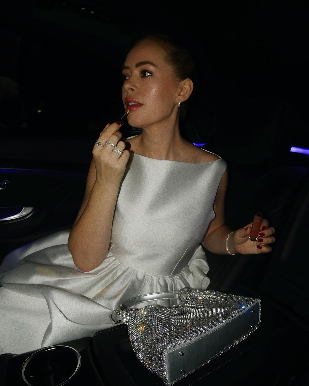 A woman in a white satin dress applies lipstick while sitting in a dimly lit car, with a sparkling silver handbag next to her.