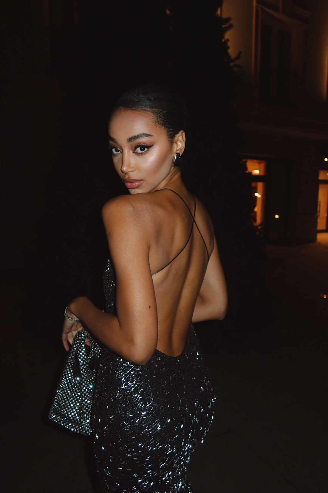 A woman in a sparkling, backless black dress with thin straps stands with her back partially turned towards the viewer, looking over her shoulder. She is holding a shimmering handbag and appears to be outdoors at night with dark surroundings.