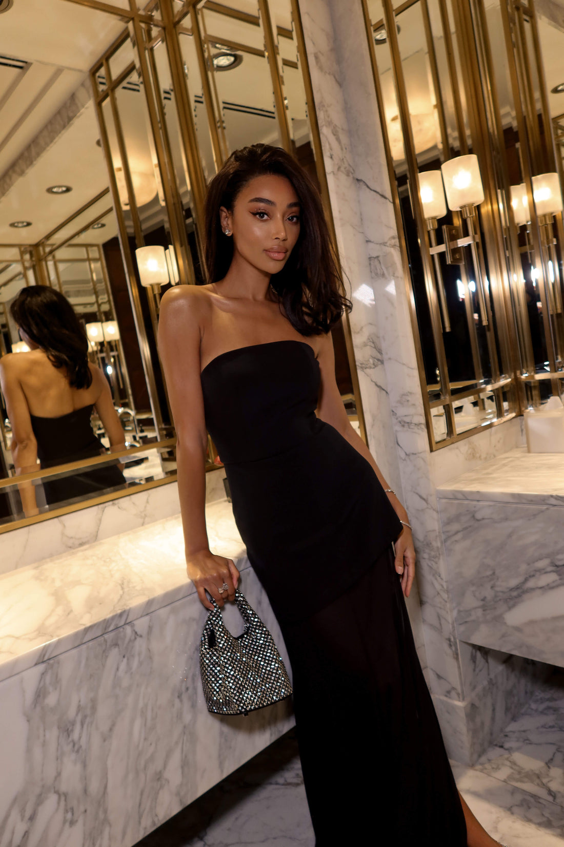 Woman in a black strapless dress holding a clutch in a marble-walled room.