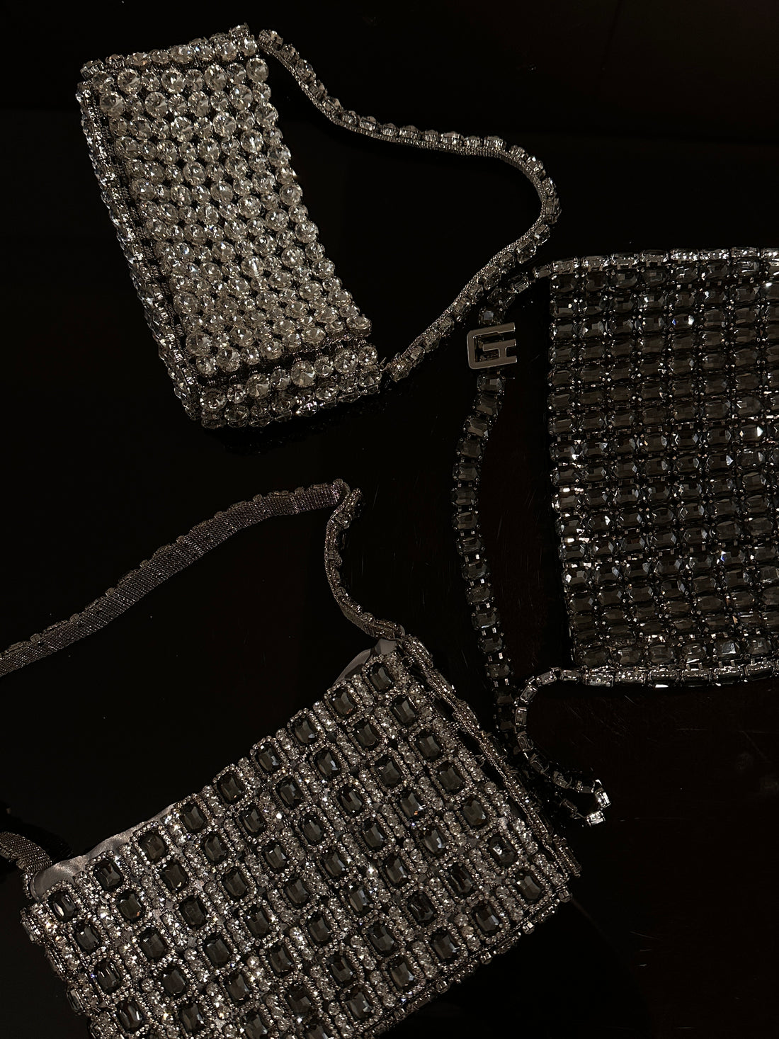 Several dark-colored, embellished bags are displayed against a black background. One bag, positioned in the upper left, is a rectangular shoulder bag covered in small, sparkling beads or crystals, with a thin, beaded strap. Another bag, in the lower right, is a rectangular clutch or shoulder bag featuring a grid pattern of larger, dark, square embellishments, also with a beaded strap. A third, partial view of a similarly embellished rectangular object is visible on the right side of the image.