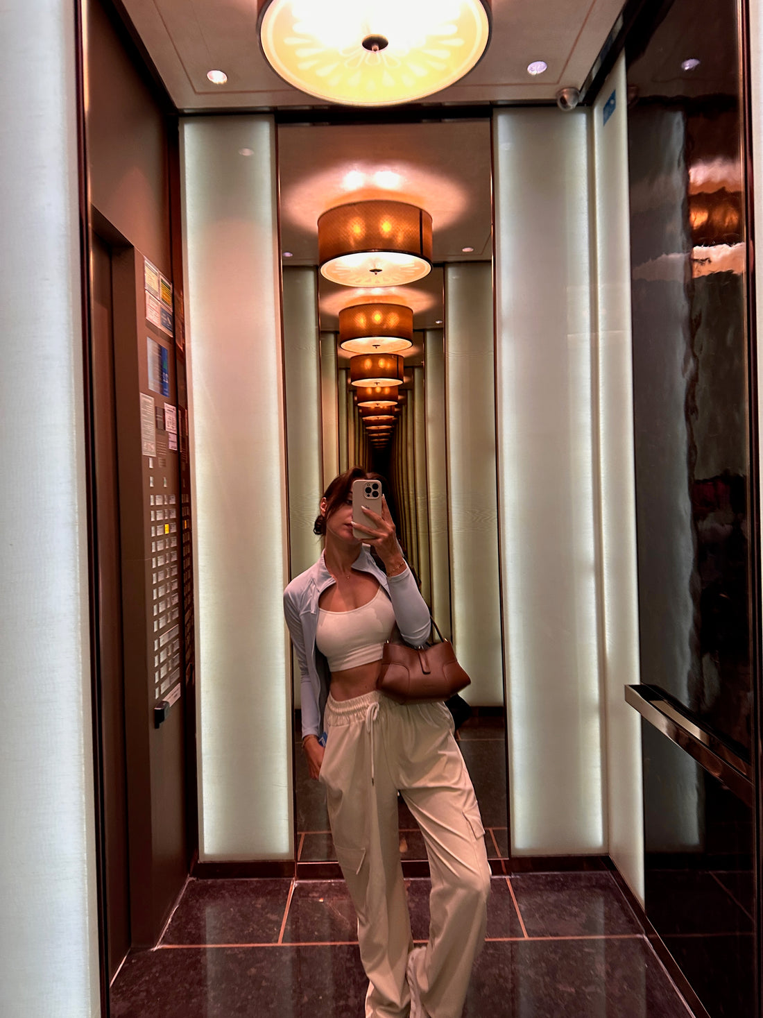 A Women wearing a white crop top, light blue collared shirt, and light-colored cargo pants takes a mirror selfie in a well-lit hallway with multiple circular ceiling lights and reflective wall panels. The person is holding a brown handbag.