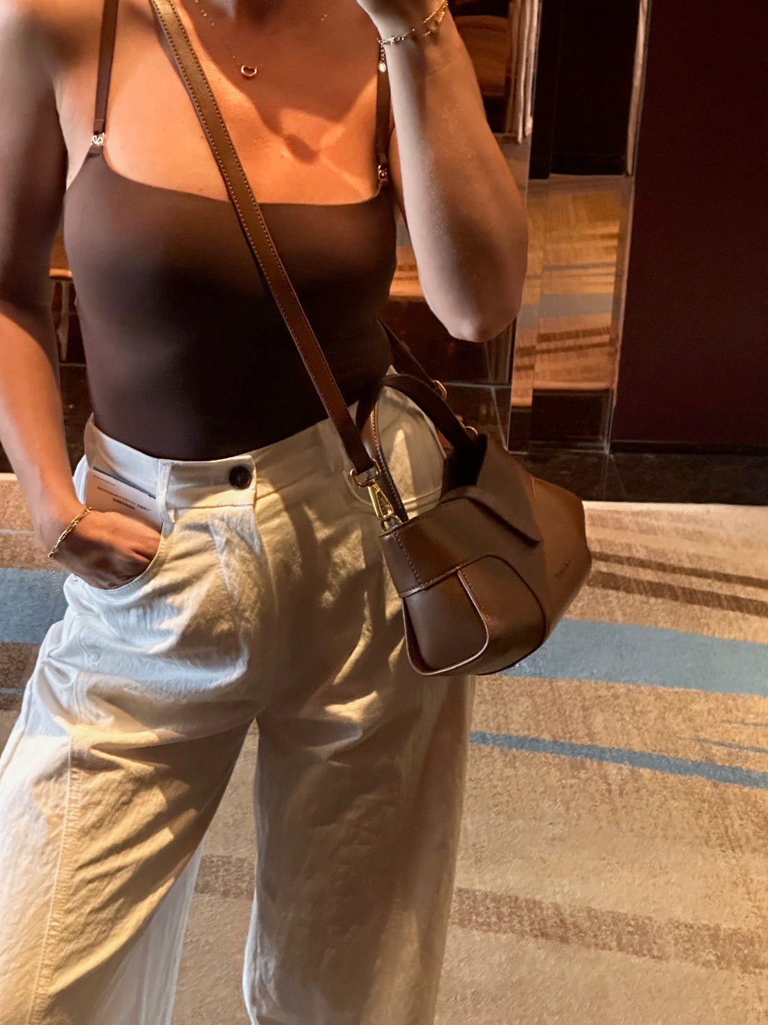 A women wearing a brown top and light-colored pants, carrying a brown shoulder bag, standing indoors.