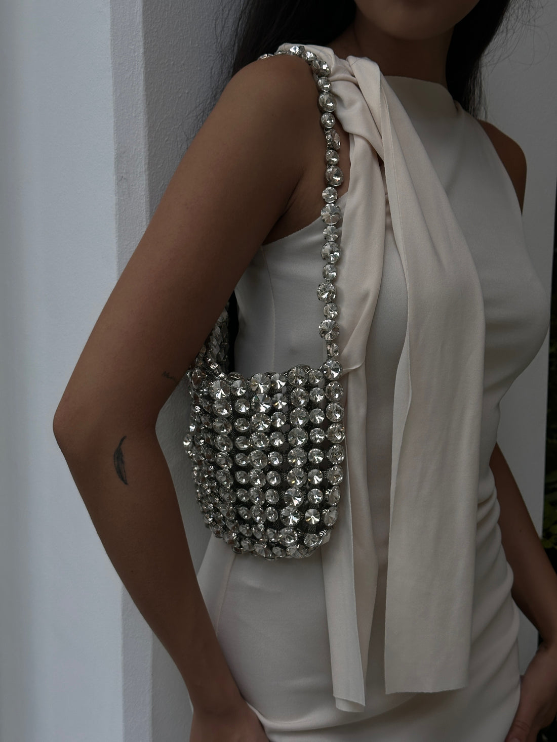 A Women wearing a light-colored, possibly white or cream, sleeveless dress or top, carrying a dazzling silver beaded or crystal-embellished handbag on their shoulder. The bag features a matching beaded strap, and the person's arm, with a small feather tattoo, is visible. The background appears to be a plain, light-colored wall or structure. 