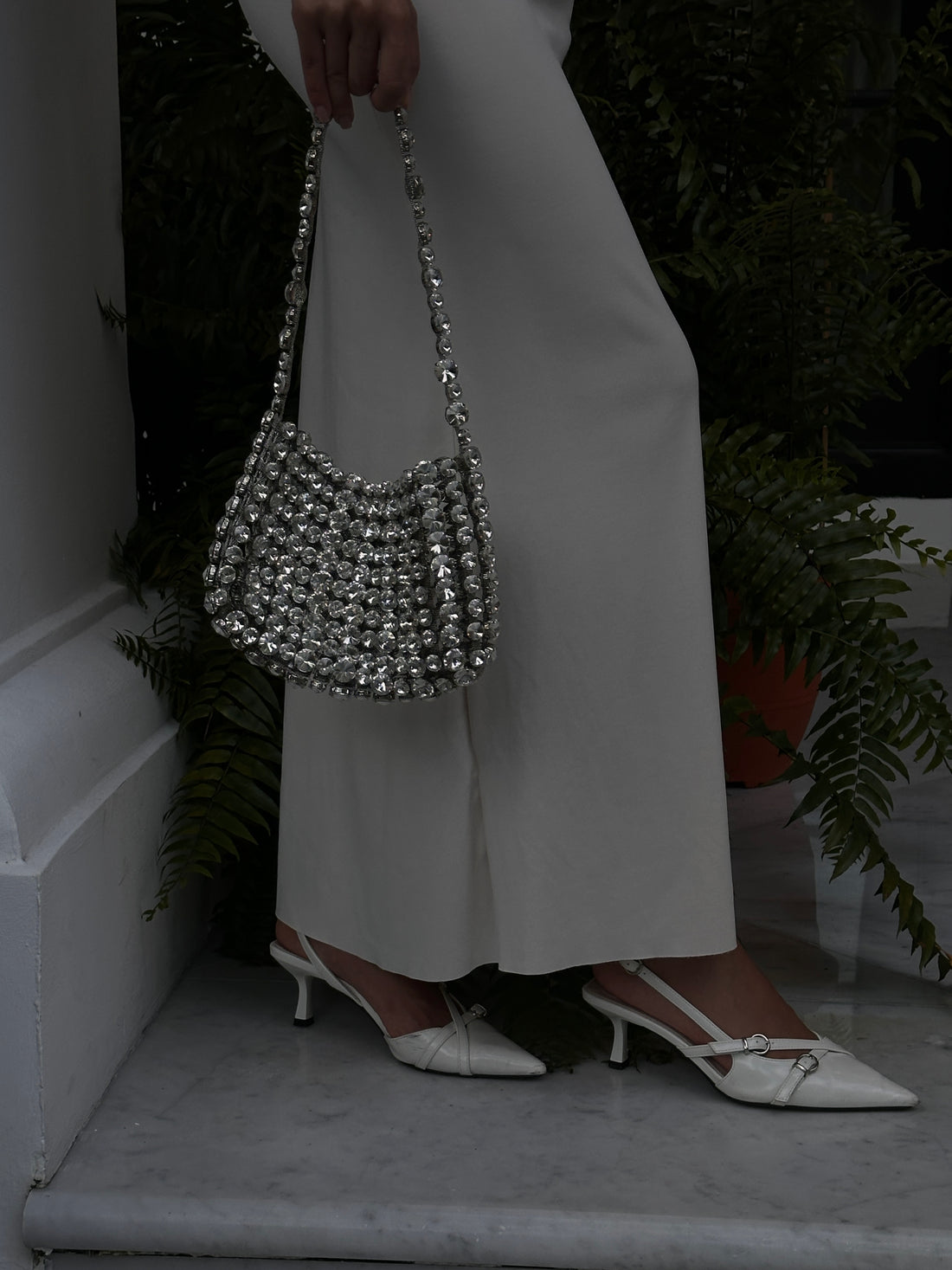 A close-up shot of a person wearing a long, light-colored dress and white slingback heels with a metallic buckle detail, carrying a shimmering, silver-toned beaded or woven handbag. The person is standing on a marbled surface next to dark foliage.