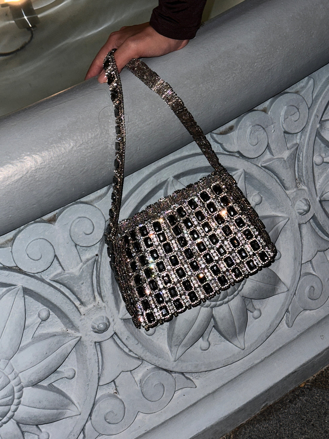 A close-up shot shows a hand holding a metallic silver shoulder bag adorned with black square beads or crystals, resting on a decorative grey stone or concrete railing. The bag features a woven metal mesh or beaded construction with a structured shape and a matching chain-link strap. The railing has intricate floral and geometric carvings, providing a textured background for the bag. 