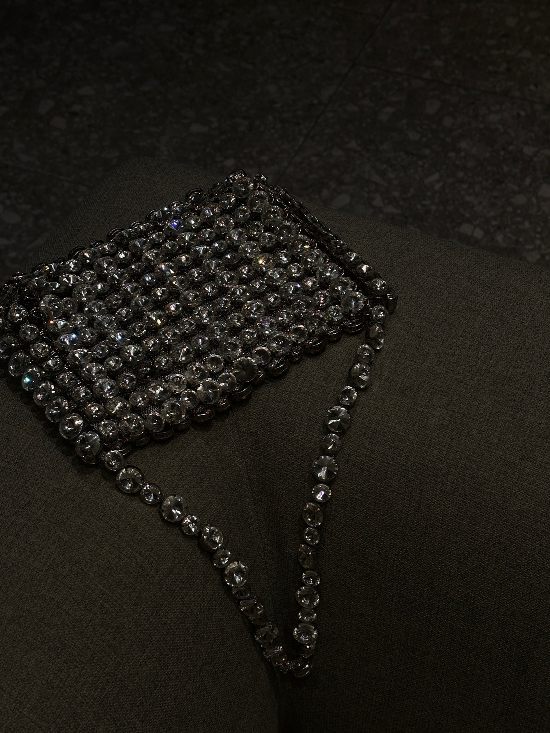 A black, rectangular object covered in shimmering, bead-like embellishments, possibly a clutch bag or a large bracelet, resting on a dark surface. The beads reflect light, creating a sparkling effect.