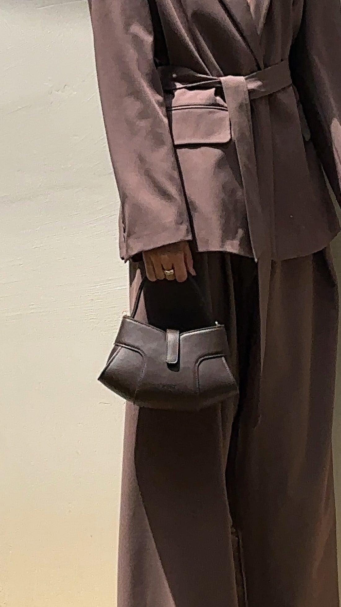 A women wearing a matching brown suit with a belted blazer and wide-leg trousers, holding a dark brown structured handbag with a unique curved flap closure.