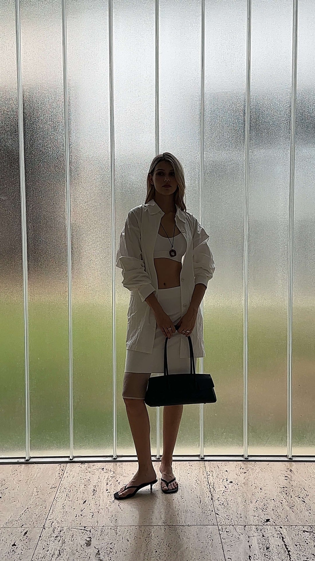 A full-shot image of a woman standing in front of a textured glass wall, holding a black handbag. She is wearing a white two-piece outfit consisting of a cropped top and a skirt, paired with dark-colored open-toed sandals. The lighting creates a silhouette effect, with the background visible as a soft green glow through the glass.