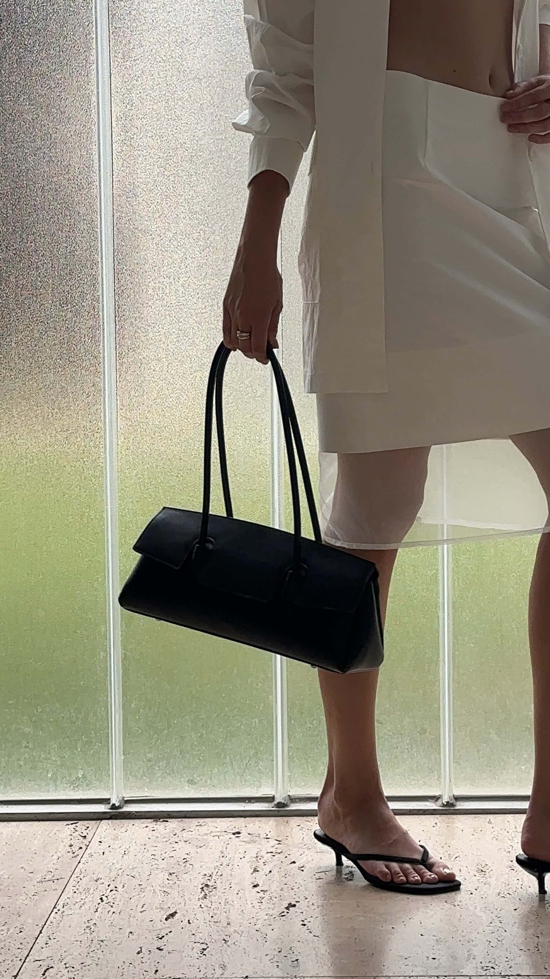 A Women wearing a white shirt and a white skirt holds a black, elongated shoulder bag in their right hand, standing on a tiled floor next to a textured glass wall with greenery visible outside. They are wearing black kitten heel flip-flops.