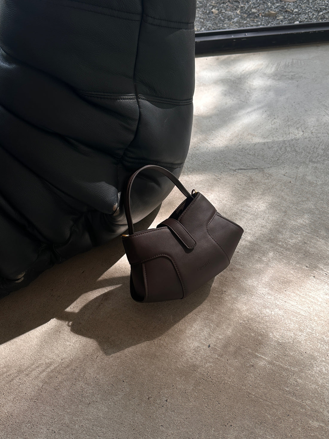 A dark brown or black small handbag with a single handle rests on a light-colored concrete or stone floor, casting a shadow. In the background, a dark, possibly leather, upholstered piece of furniture is partially visible.