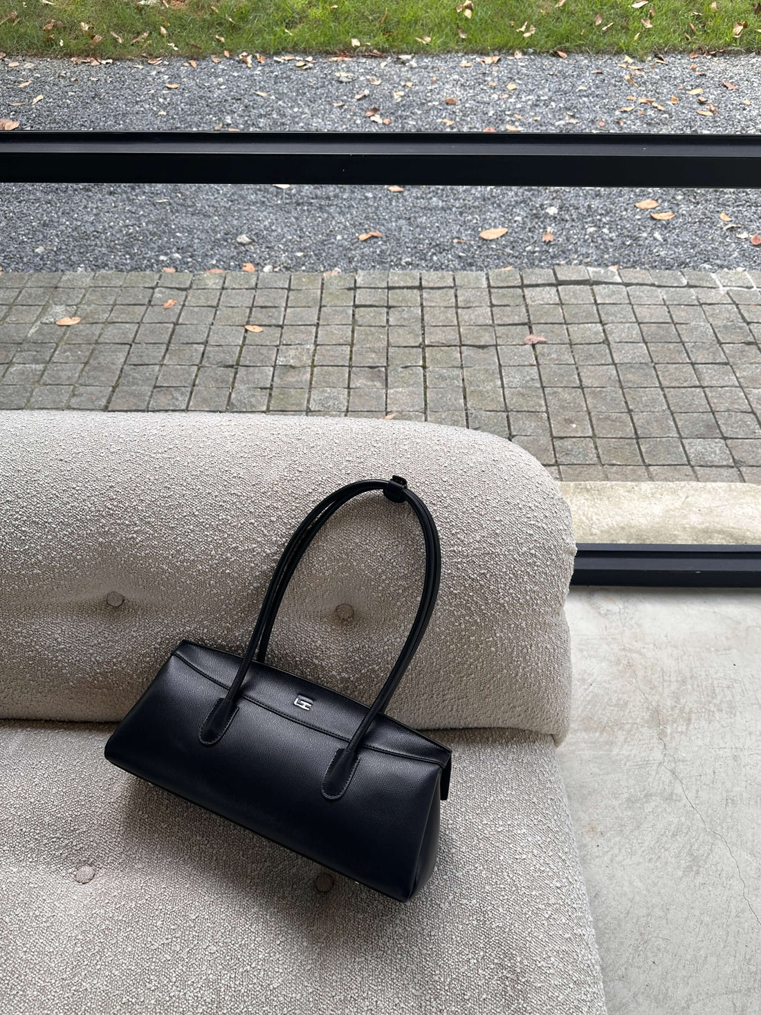 A black handbag with a top handle rests on a textured, light-colored sofa, positioned in front of a large window overlooking an outdoor area with gravel and green foliage.