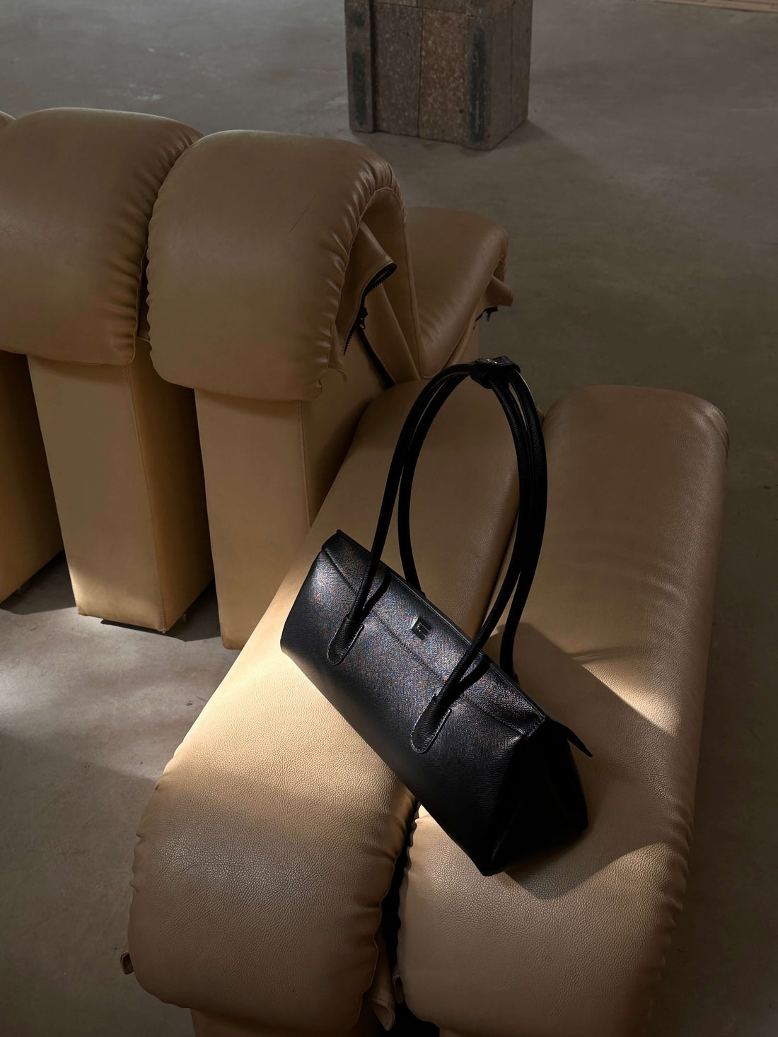 A black cylindrical handbag with a long handle rests on a light-colored, textured armchair in a dimly lit room, with a beige, padded sofa visible in the background.