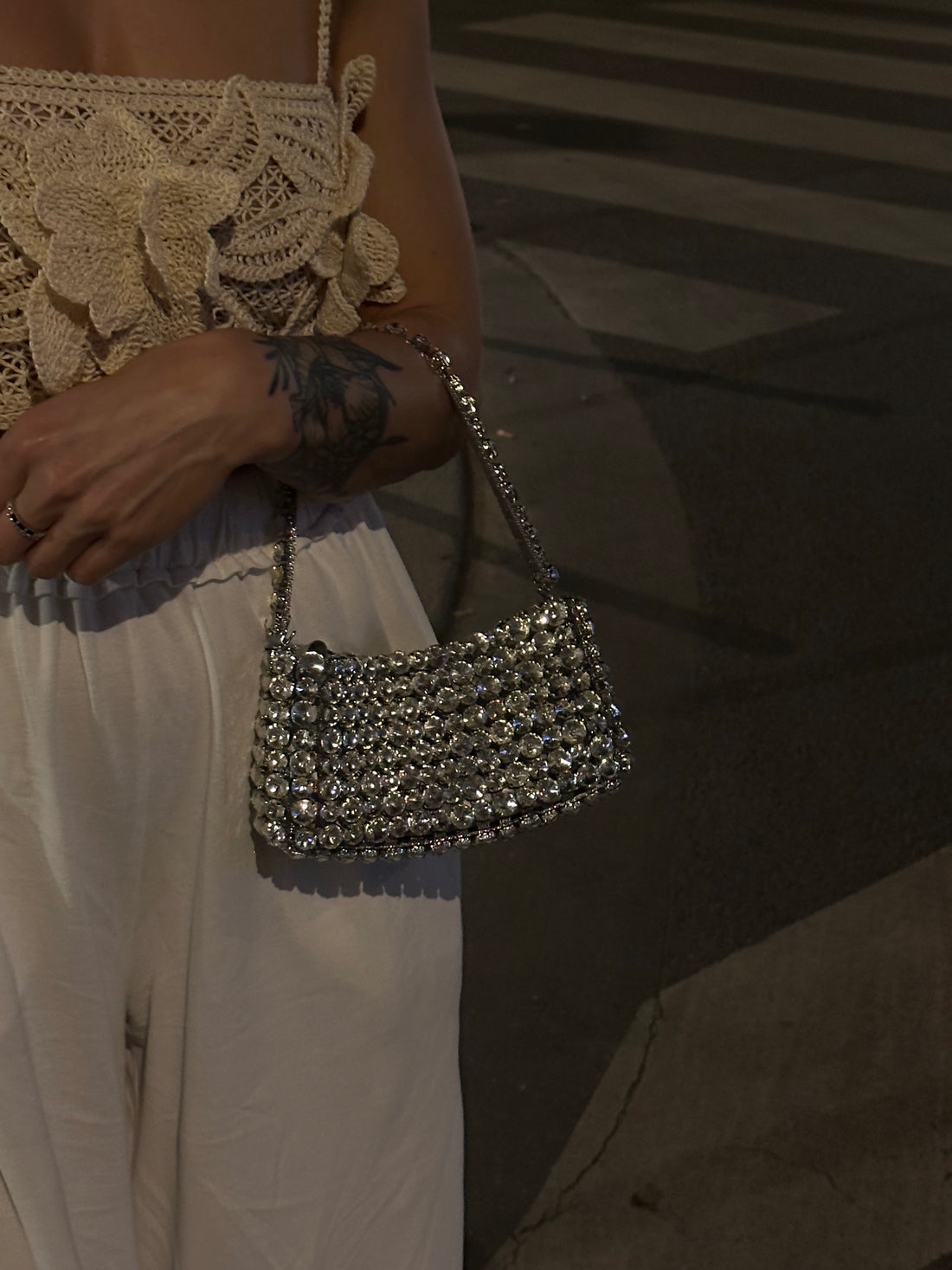 a person wearing a white crocheted top and white pants, holding a shimmering, beaded or rhinestone-covered handbag with a short strap. The background is dark and appears to be an outdoor setting at night, possibly a street or sidewalk.