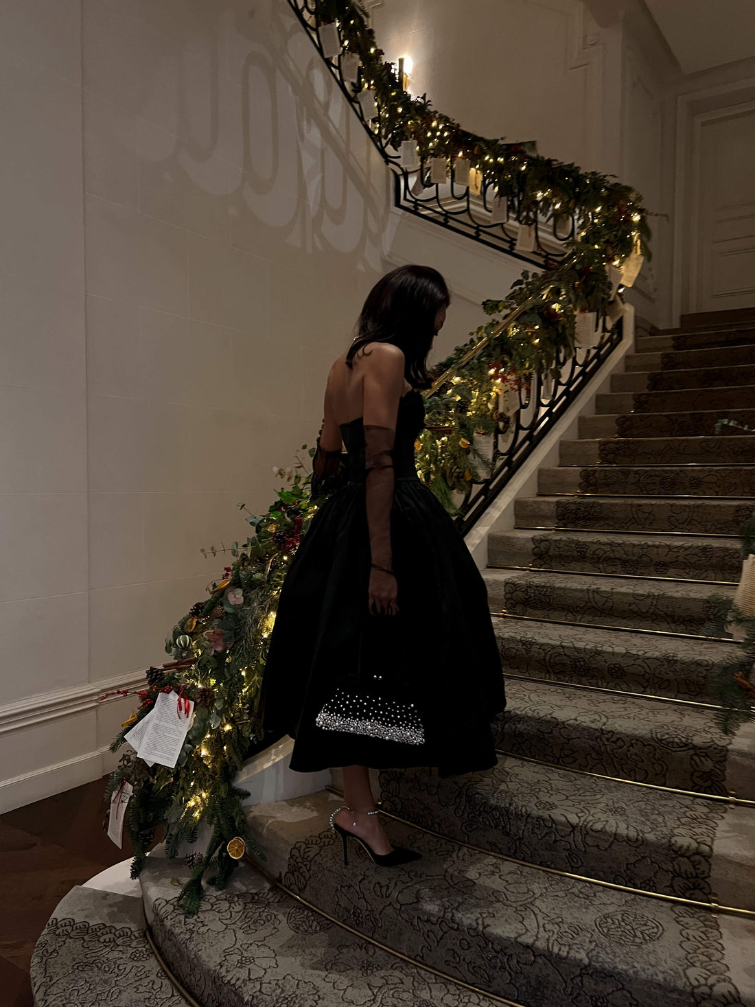 A woman in a black strapless dress and long gloves descends a grand staircase adorned with festive green garlands and lights. She holds a sparkling clutch bag and wears black high heels. The staircase has a dark railing and carpeted steps.