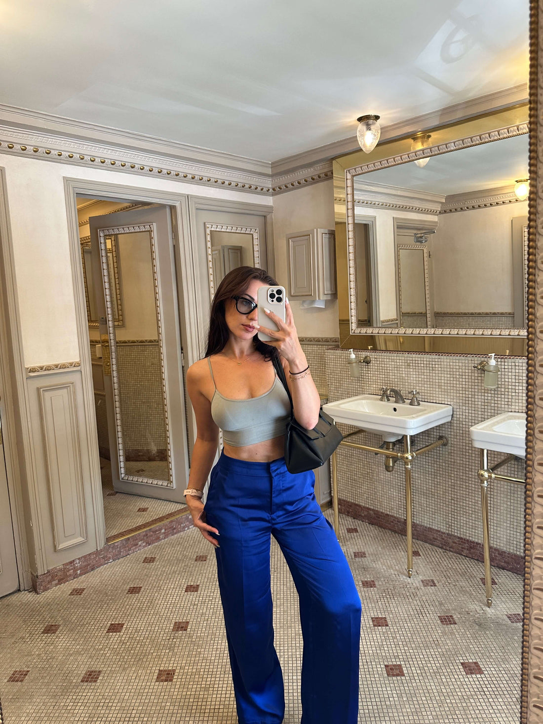 A woman with long dark hair and sunglasses takes a mirror selfie in a bathroom. She is wearing a light blue or grey crop top with thin straps and loose-fitting, shiny royal blue pants. She is holding a dark handbag and her phone, which has a prominent camera lens visible. The bathroom features ornate detailing on the walls, a large mirror with a gilded frame, and white pedestal sinks with brass fixtures. The floor is tiled with a patterned design.
