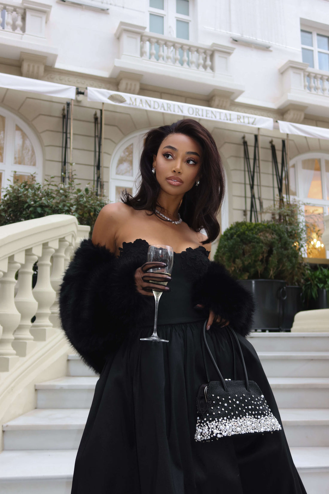 A woman in a black off-the-shoulder gown with a fur stole and a beaded handbag stands on steps in front of a building labeled MANDARIN ORIENTAL RITZ. She holds a wine glass in one hand.