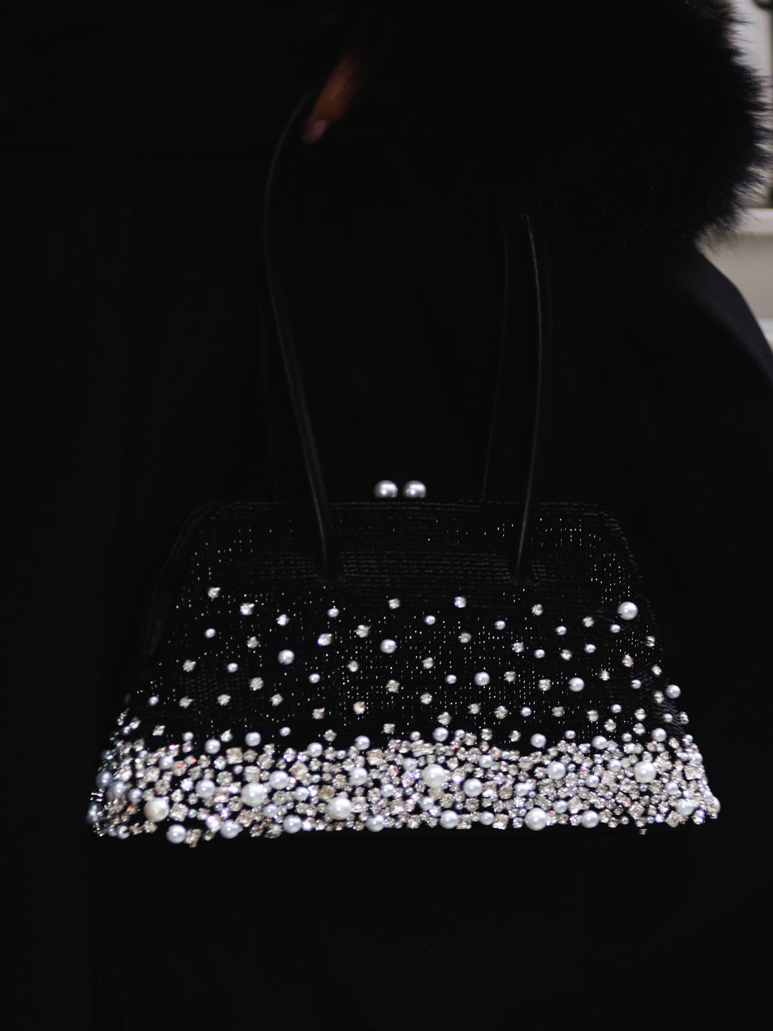 A black clutch bag, heavily embellished with cascading pearls and crystals, held against a dark background, likely a person wearing dark clothing. The bag features a structured silhouette and a top clasp with two pearl-like embellishments. 
