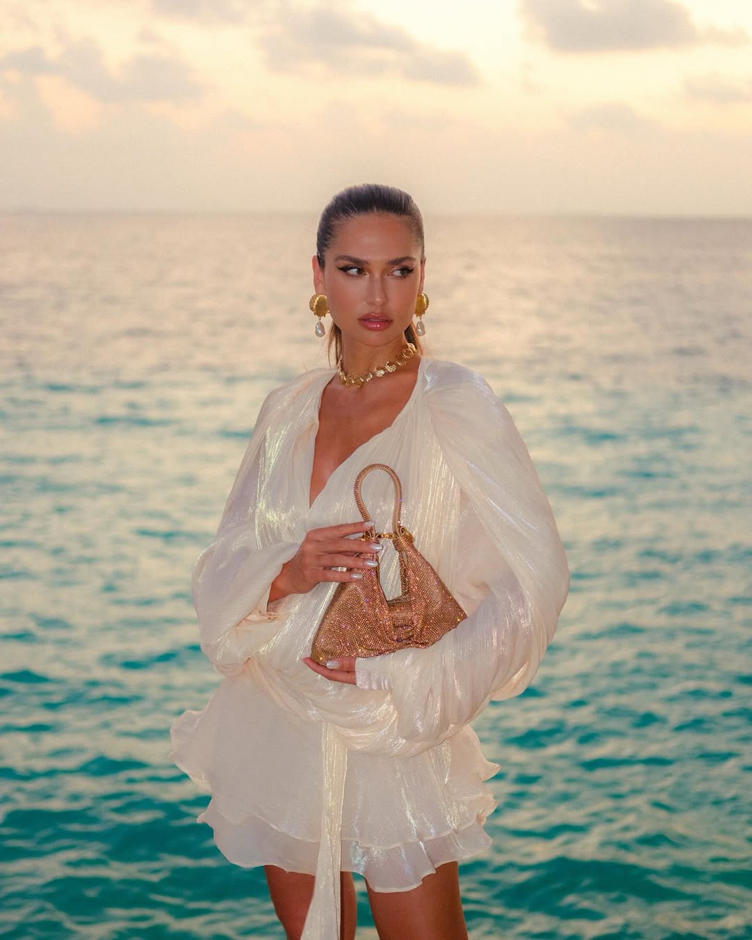 A woman with her hair pulled back stands in front of a body of water at sunset, holding a small, woven straw-colored handbag. She is wearing a flowing, sheer white dress or cover-up with voluminous sleeves and a deep V-neckline, along with gold-colored earrings and a choker necklace. The background shows the ocean with a soft, warm glow on the horizon, suggesting either sunrise or sunset.