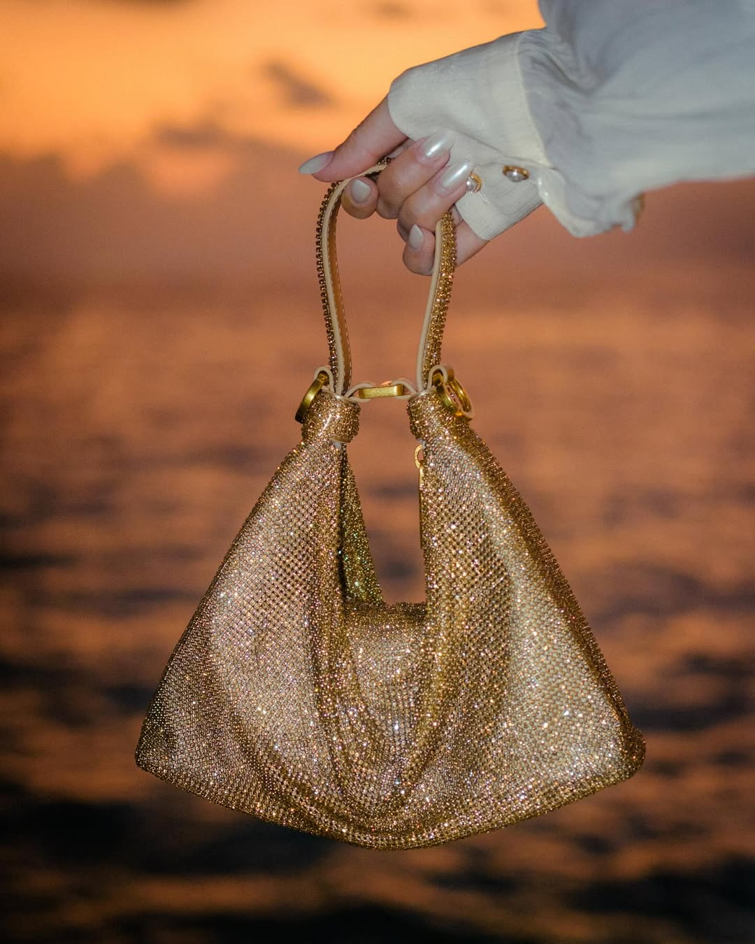 A hand holds a shimmering gold rhinestone or sequin evening bag against a blurry background of a sunset over water. The bag is made of a mesh-like material covered in small, reflective elements, and has a braided strap and gold hardware. The hand has manicured nails.