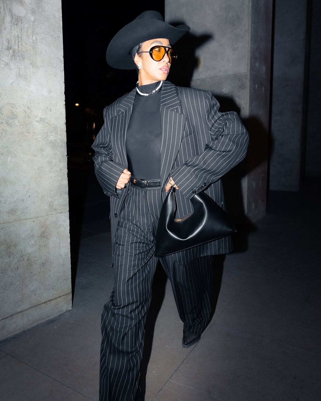 Alt text for this image: A person wearing a black pinstripe oversized suit, a black turtleneck, a black cowboy hat, and orange-tinted sunglasses, carrying a black hobo-style handbag, walks confidently on a dimly lit street or pathway.
