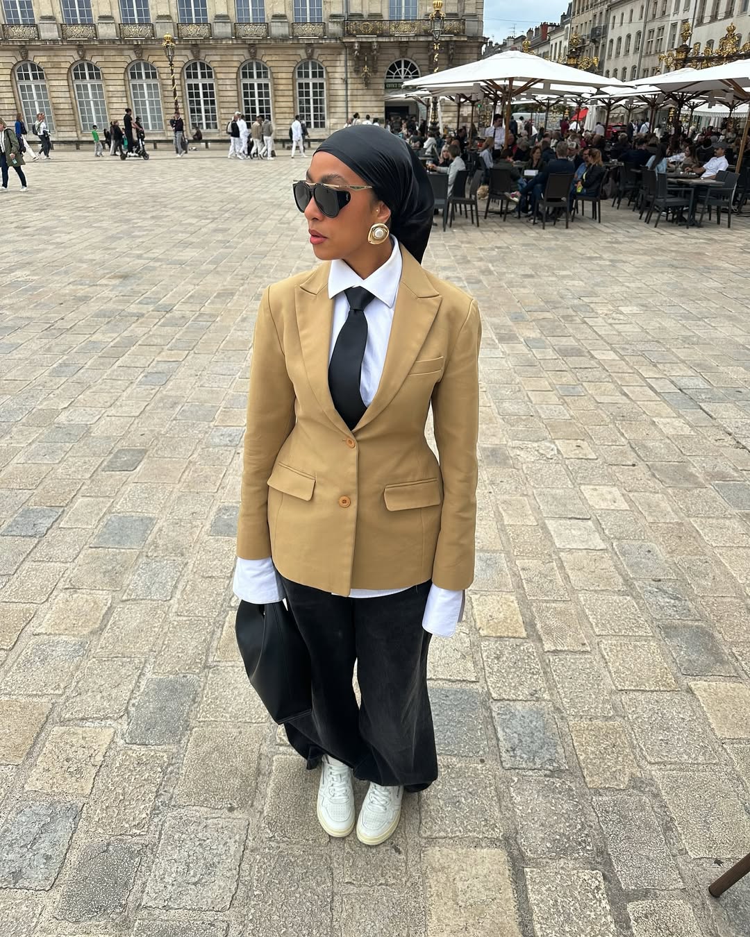A person wearing a tan blazer, white shirt, black tie, black headscarf, black wide-leg pants, and white sneakers stands on a cobblestone plaza, looking to the right. They are also wearing sunglasses and large pearl earrings. In the background, there is a building with arched windows and a cafe area with tables and umbrellas.