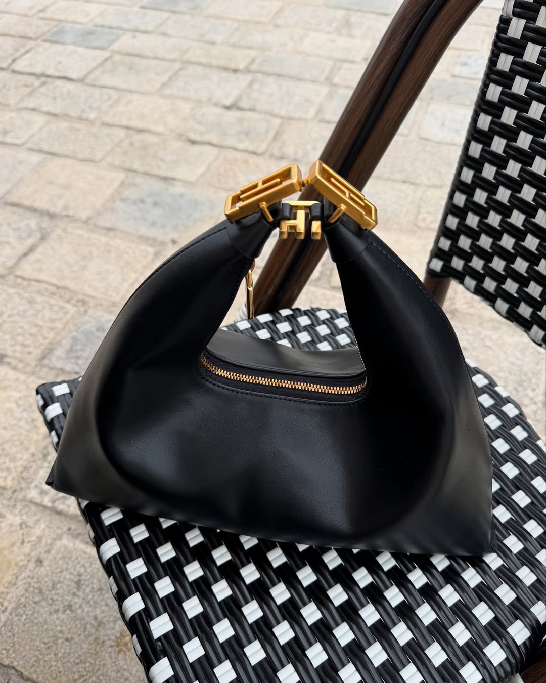 A black leather handbag with a unique triangular shape and gold-toned hardware on the handle, resting on a black and white checkered chair outdoors.