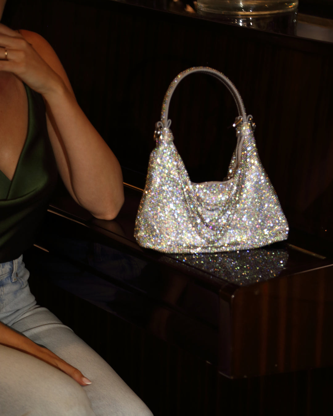 A close-up shot of a sparkling silver rhinestone handbag, identified as the Jules bag by Giarite, resting on a dark, polished surface, likely a piano, with a person's arm and leg visible in the background. 