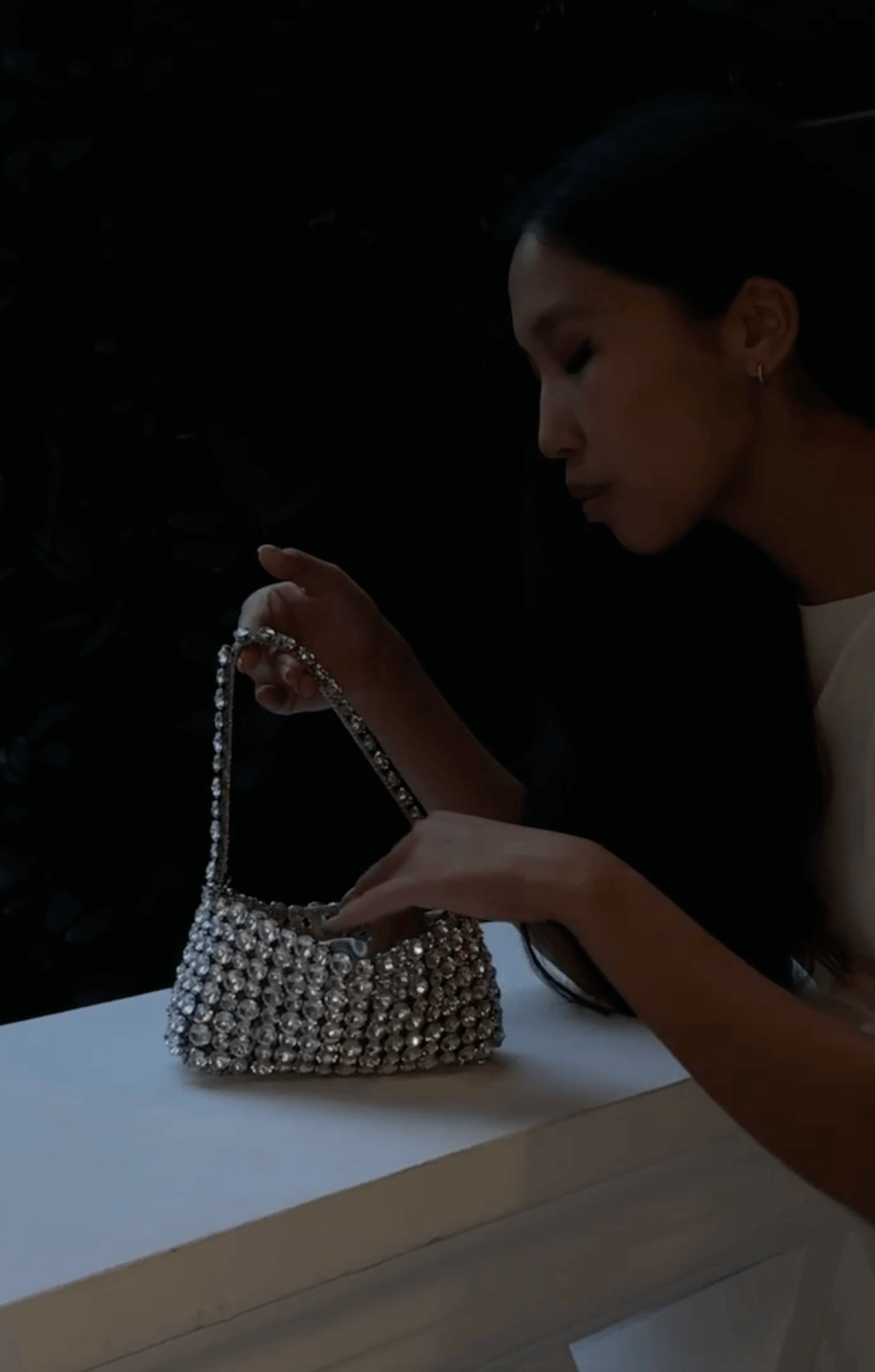 A woman in a dark setting holds a shimmering, silver beaded or crystal-embellished handbag with a top handle, resting it on a white surface.