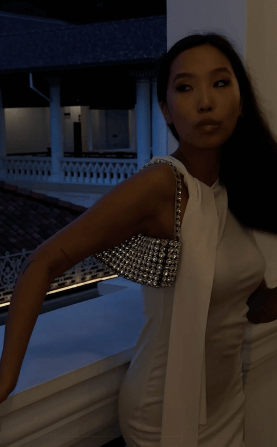 A woman in a white dress and a sparkly shoulder bag stands on a balcony at dusk or night. The background shows a building with arches and a tiled roof, dimly lit.