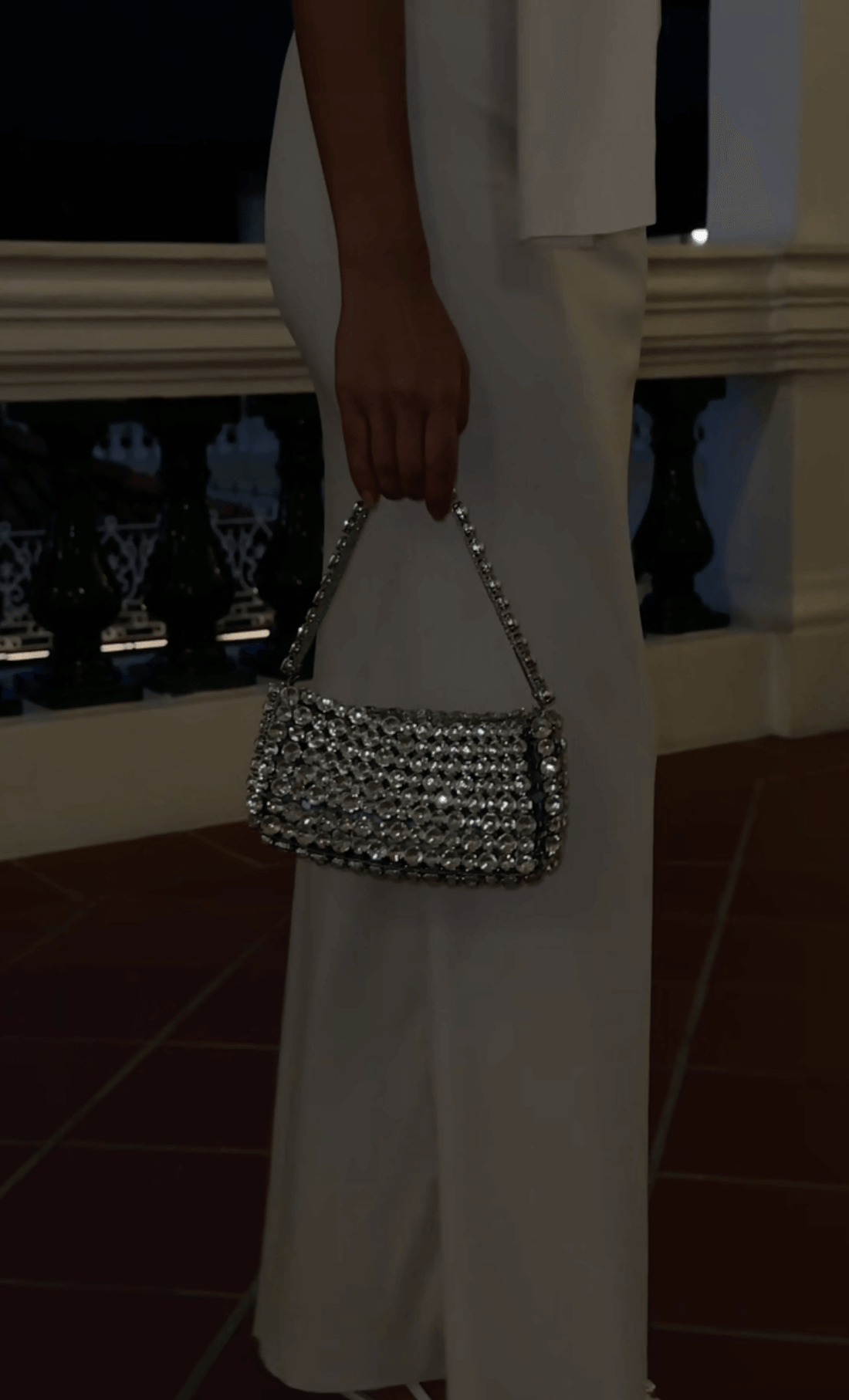  A Women in a white dress holding a rectangular, silver, rhinestone-embellished handbag with a matching handle. The background is dimly lit, revealing a dark railing and a hint of architectural details.