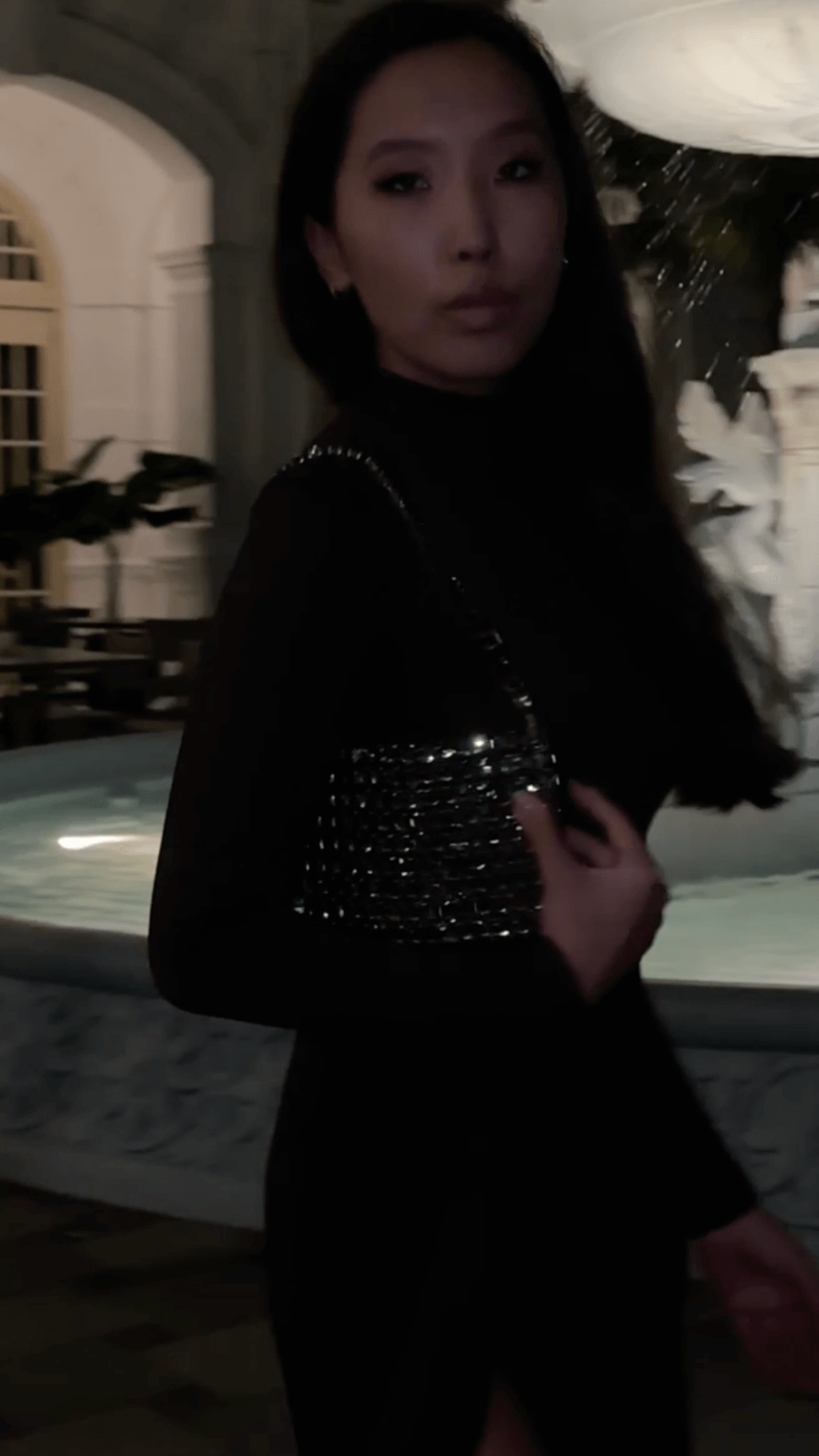 A young woman with long dark hair, wearing a black long-sleeved dress and carrying a sparkling black shoulder bag, stands outdoors at night near a stone fountain with a statue. The background features architectural elements like an archway and a building with windows, suggesting an elegant or historic setting.