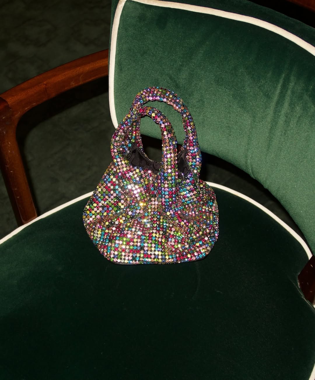 A vibrant, multicolor crystal-embellished mini handbag with a knotted handle, placed on a dark green velvet chair with white piping. 