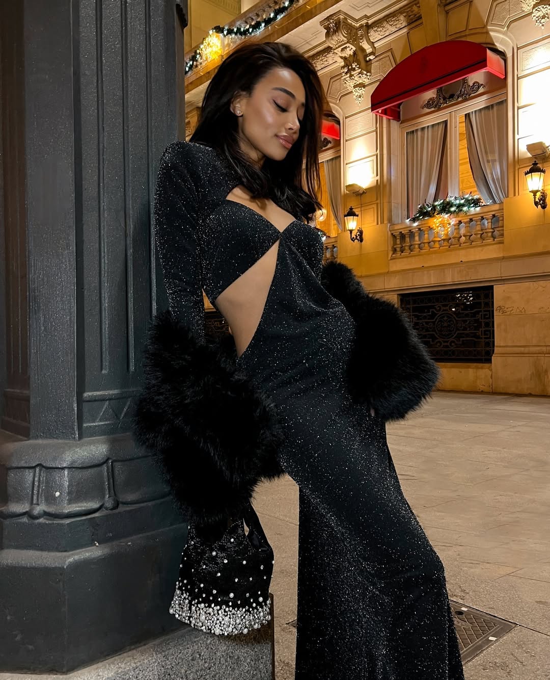 A woman with dark hair wearing a black glittery cutout dress with a fur stole and a beaded handbag, posing outdoors at night with a building in the background.
