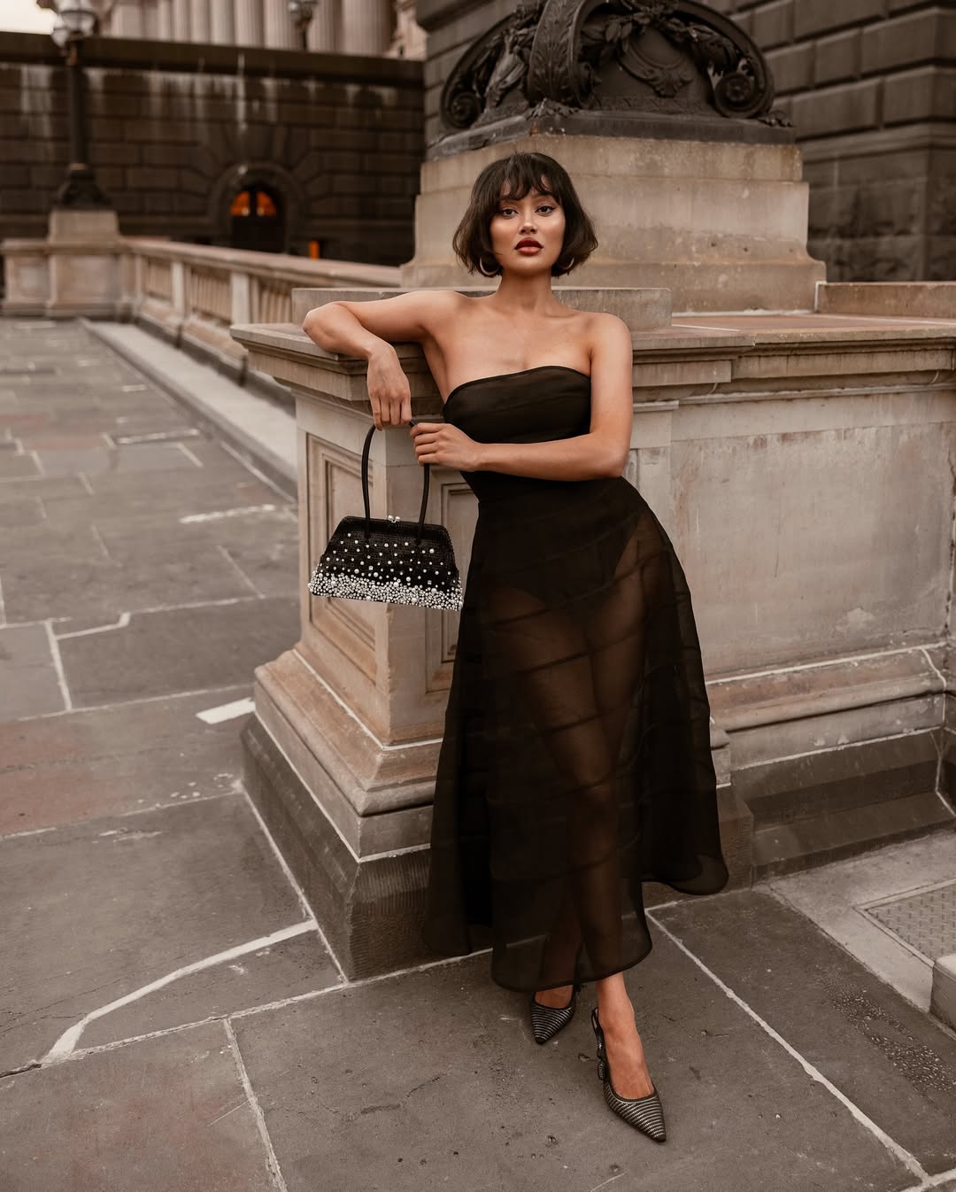 A woman with a bob haircut and a black strapless sheer midi dress stands outdoors, leaning on a stone railing while holding a black embellished handbag.
