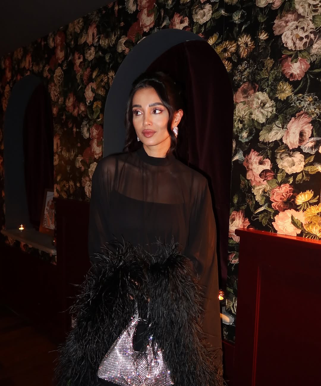 A woman in a black sheer top and a black feathered coat holds a sparkly silver handbag while standing in front of a dark floral wallpaper.