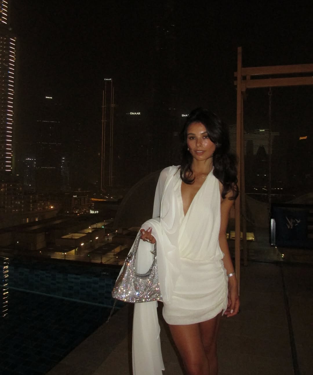 A woman in a white dress and carrying a sparkling handbag stands on a rooftop at night, with a city skyline and pool visible in the background.